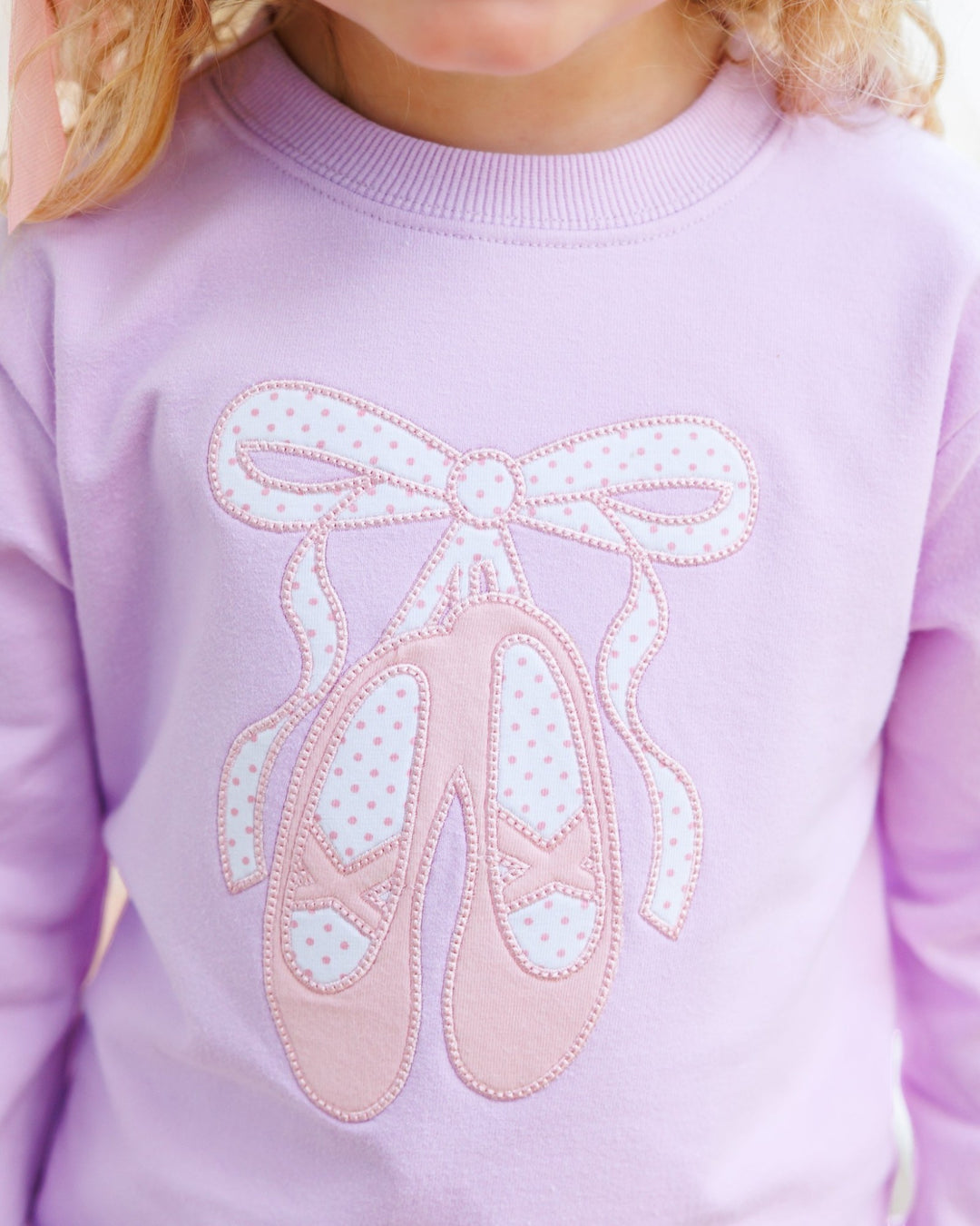 A close-up of a child wearing a light purple sweatshirt with a pink and white polka-dot ballet shoe applique on the front, that store