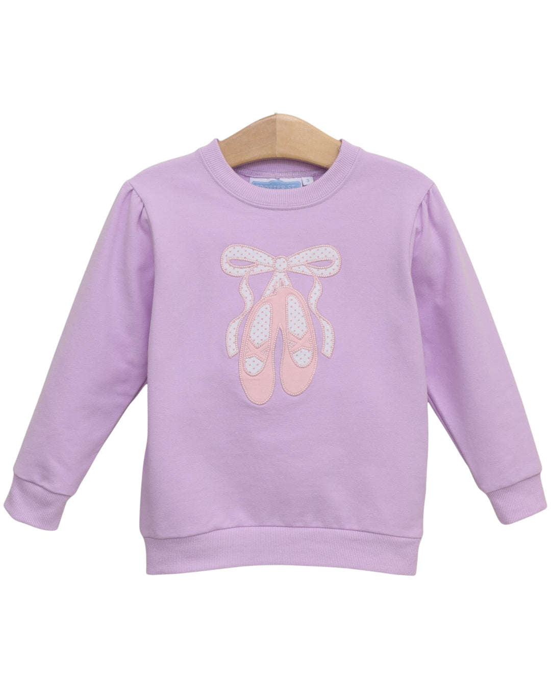 A lavender purple long-sleeve sweatshirt for kids, featuring a graphic of pink ballet slippers with white bows on the front, hanging on a wooden hanger against a white background, that store