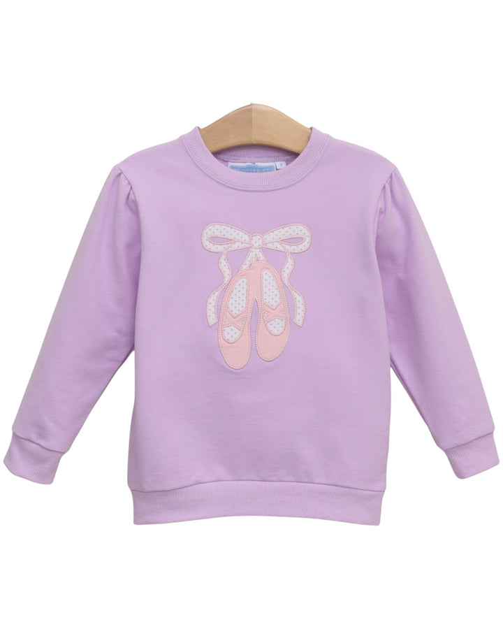 A lavender purple long-sleeve sweatshirt for kids, featuring a graphic of pink ballet slippers with white bows on the front, hanging on a wooden hanger against a white background, that store