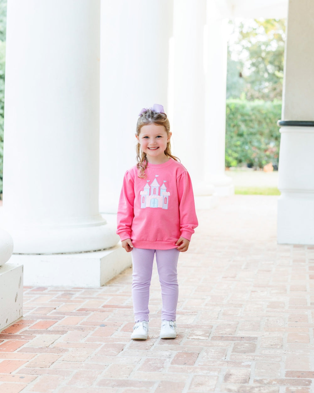 A young girl stands on a brick walkway between white columns, wearing a bright pink sweatshirt with a white castle graphic, lavender leggings, and white sneakers, that store
