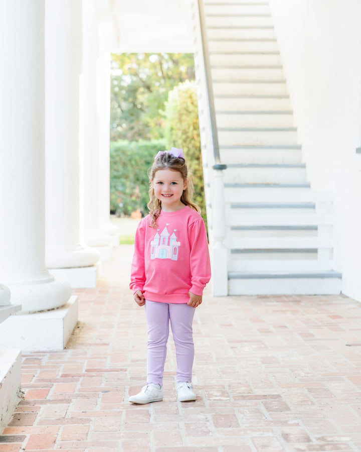 A young girl stands on a brick porch wearing a bright pink long-sleeve sweatshirt with a white castle graphic and light purple leggings, that store