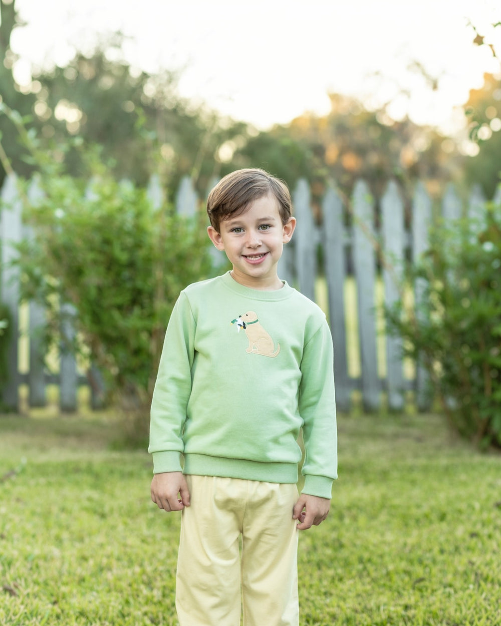 A young boy standing outdoors on a grassy lawn in front of a white picket fence, wearing a light green crewneck sweatshirt with a small dog graphic on the chest and pale yellow pants, that store