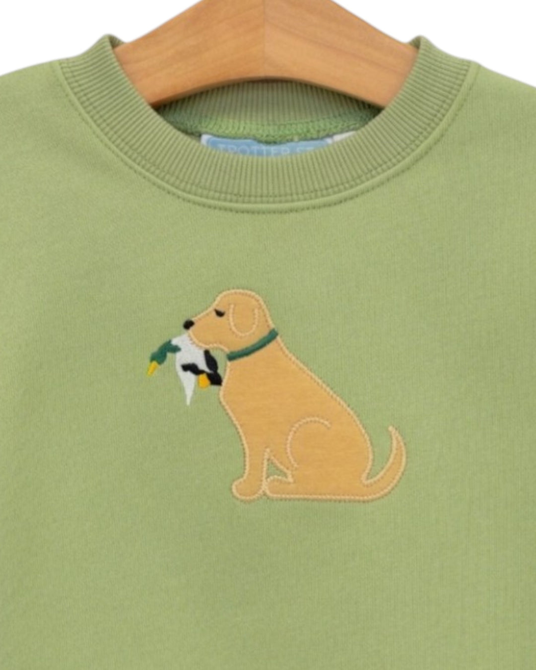 A close-up of a sage green crewneck sweatshirt on a wooden hanger, featuring an embroidered golden retriever dog holding a duck in its mouth, that store