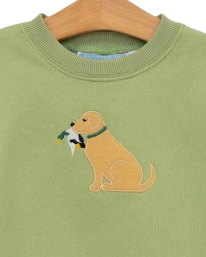 A close-up of a sage green crewneck sweatshirt on a wooden hanger, featuring an embroidered golden retriever dog holding a duck in its mouth, that store