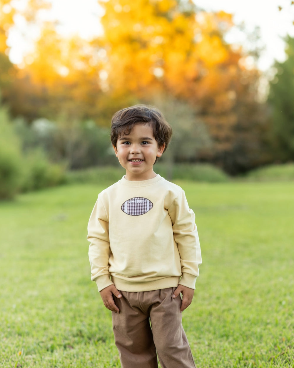 A young child standing outdoors on a grassy lawn wearing a cream sweatshirt with a football graphic and brown pants, with a blurred background of green grass and warm autumn trees, that store