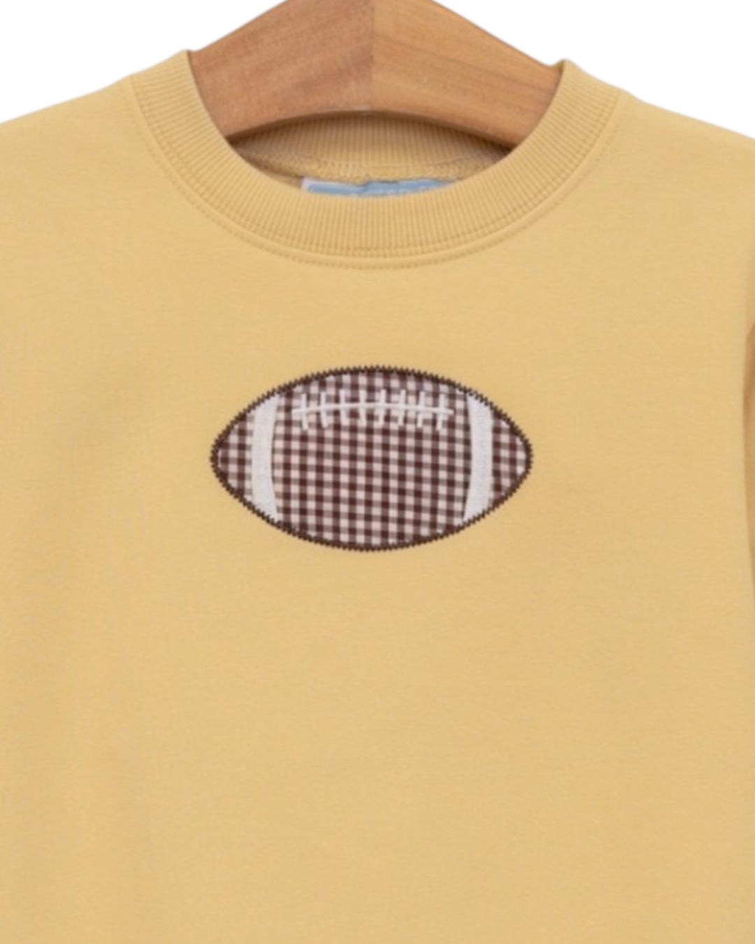 A close-up of a pale yellow crewneck sweatshirt hanging on a wooden hanger, featuring a brown and white gingham football patch with white laces on the chest, that store