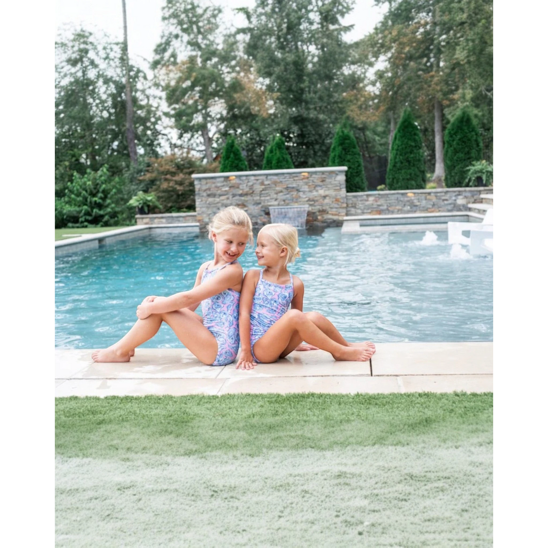 Two young girls in swimsuits sitting by a pool with trees in the background