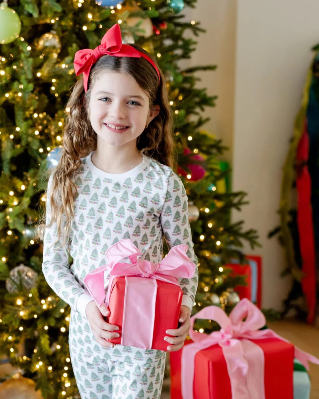 Young girl holding a gift box with a pink bow in front of a decorated Christmas tree.