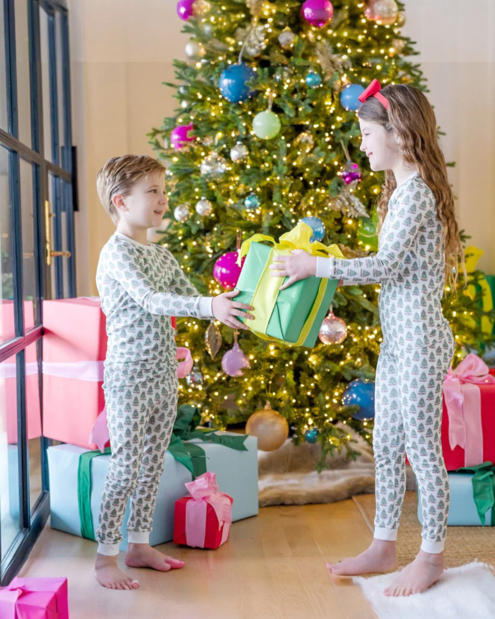 Two children in matching pajamas exchanging gifts in front of a decorated Christmas tree.
