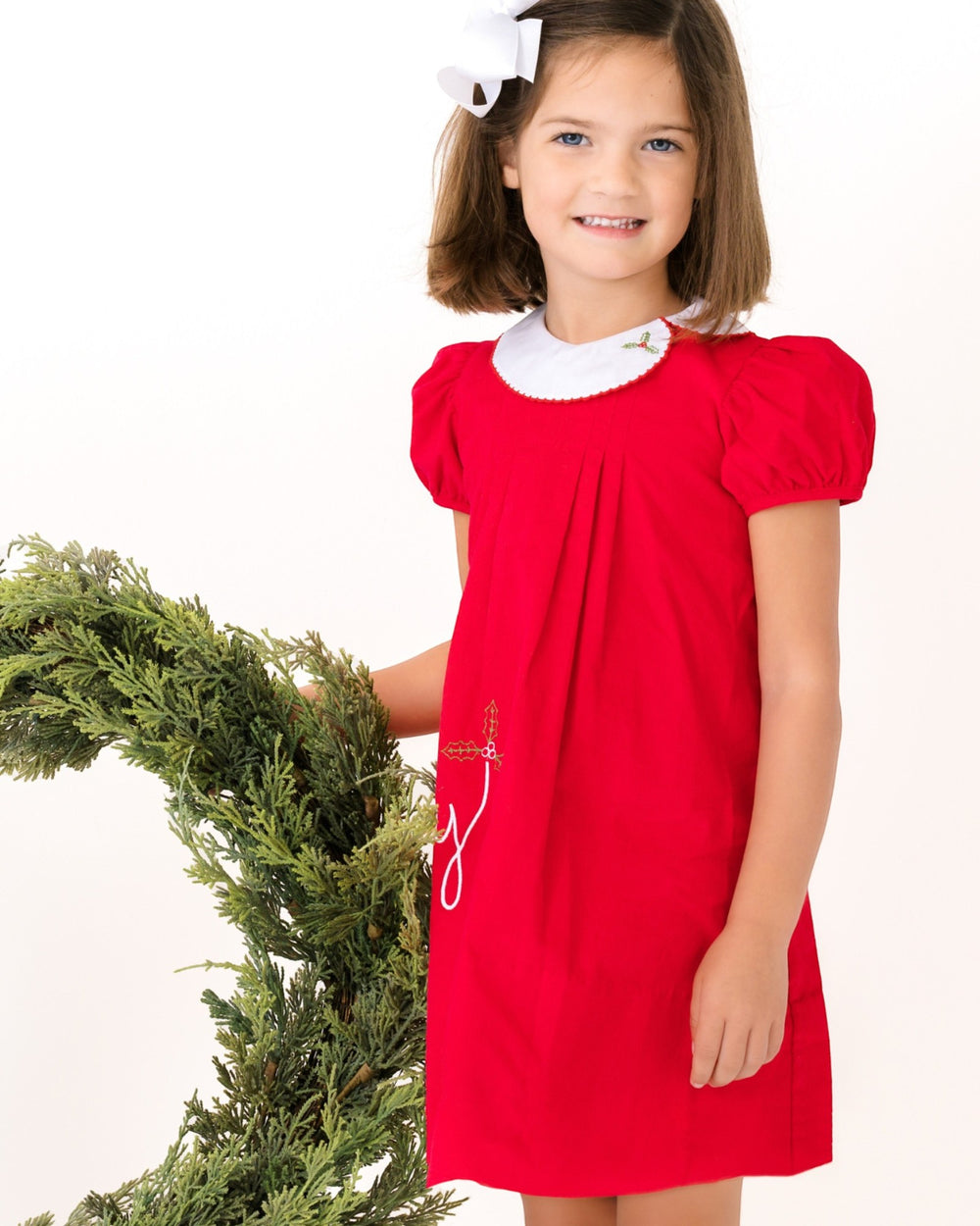 Young girl in a red dress with white collar holding a Christmas wreath on a white background