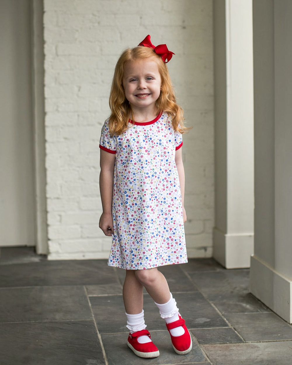 Young girl in a floral dress with red trim and shoes, standing indoors.