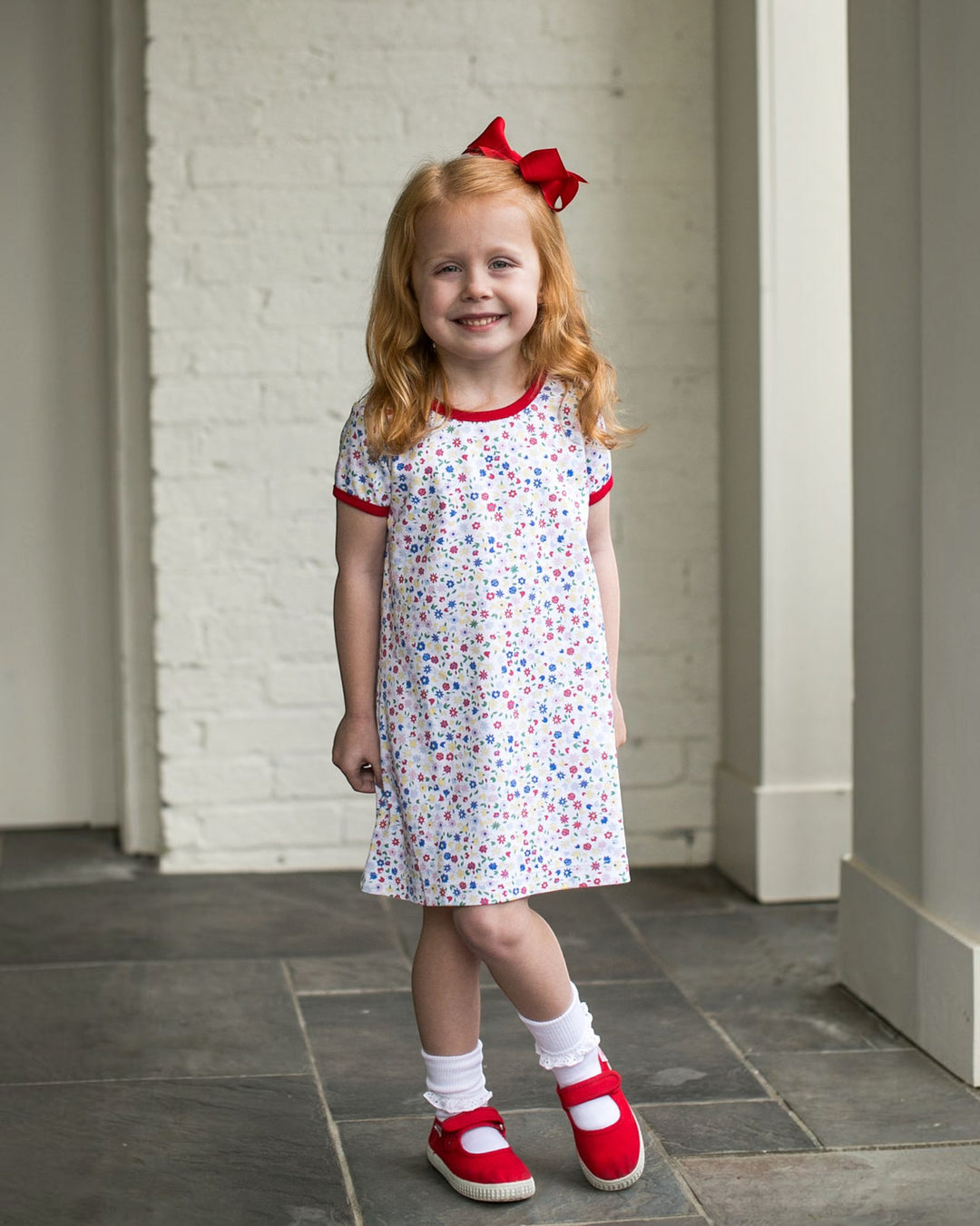 Young girl in a floral dress with red trim and shoes, standing indoors.