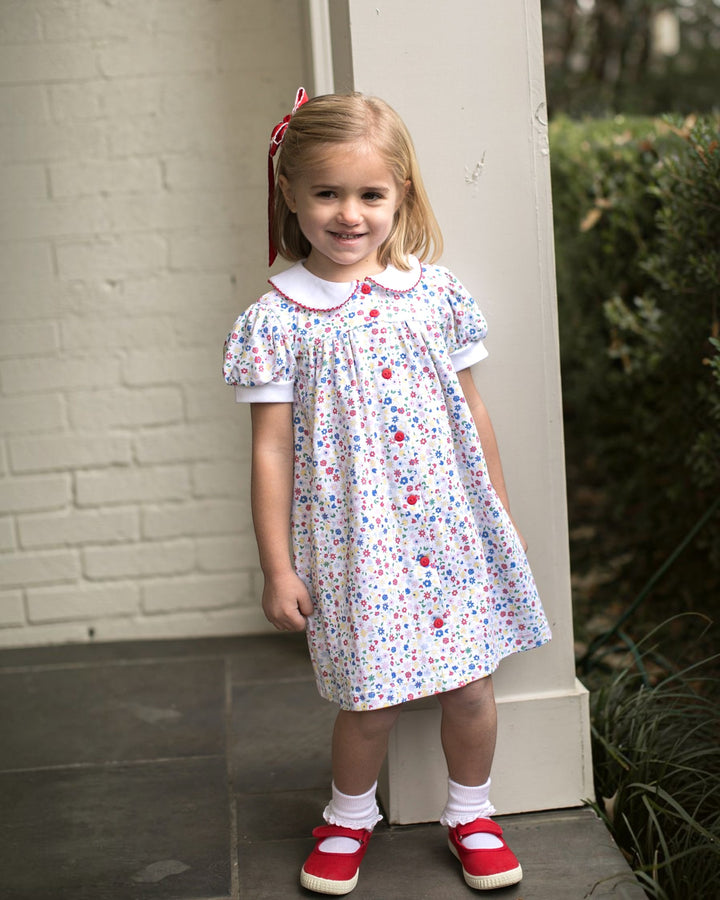 Young girl in a floral dress standing against a brick wall.