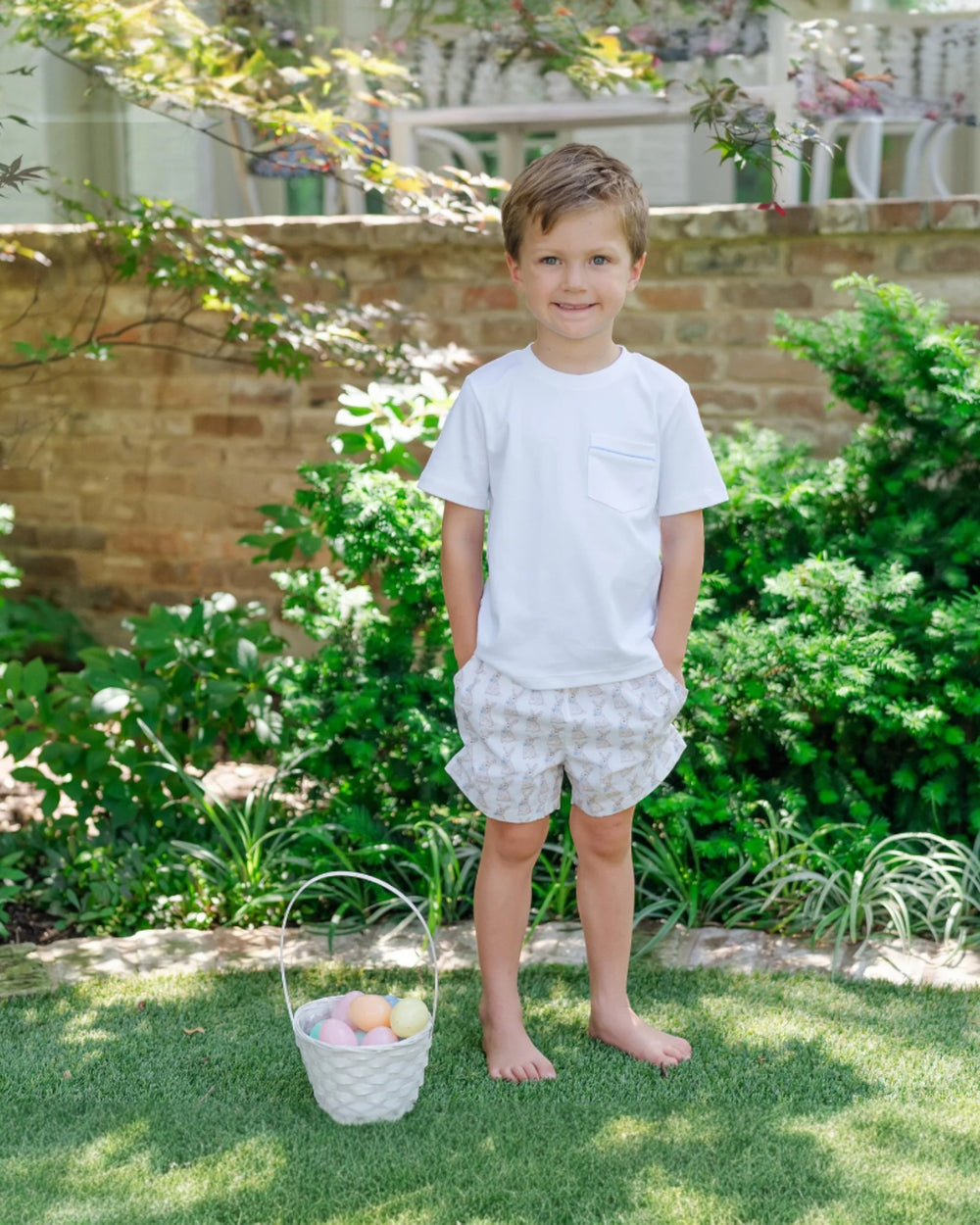 Young boy standing outdoors with a basket of Easter eggs, surrounded by greenery.