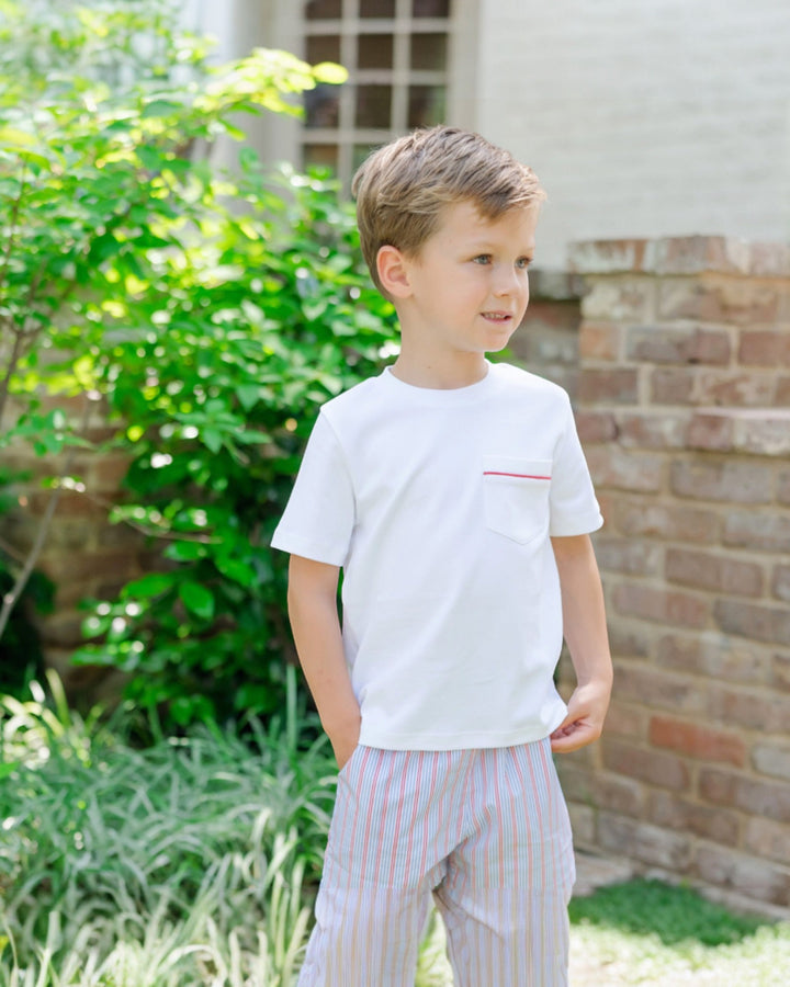 Young boy wearing a white t-shirt with a red stripe and striped pants standing outdoors.