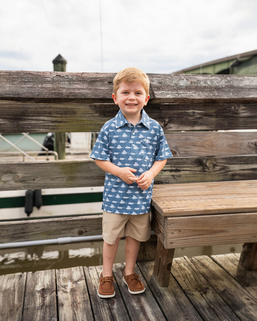 Young boy standing on a wooden dock wearing a blue patterned shirt and beige shorts.