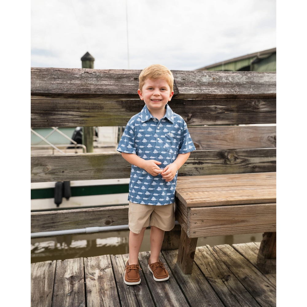 Young boy standing on a wooden dock wearing a blue patterned shirt and beige shorts.
