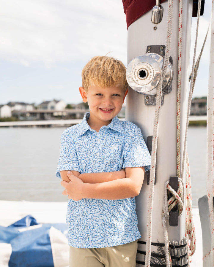 Young boy in a blue patterned shirt standing on a boat with water and sky in the background