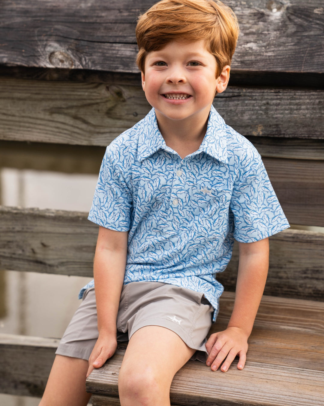 Young boy sitting on a wooden bench wearing a blue patterned shirt and gray shorts.
