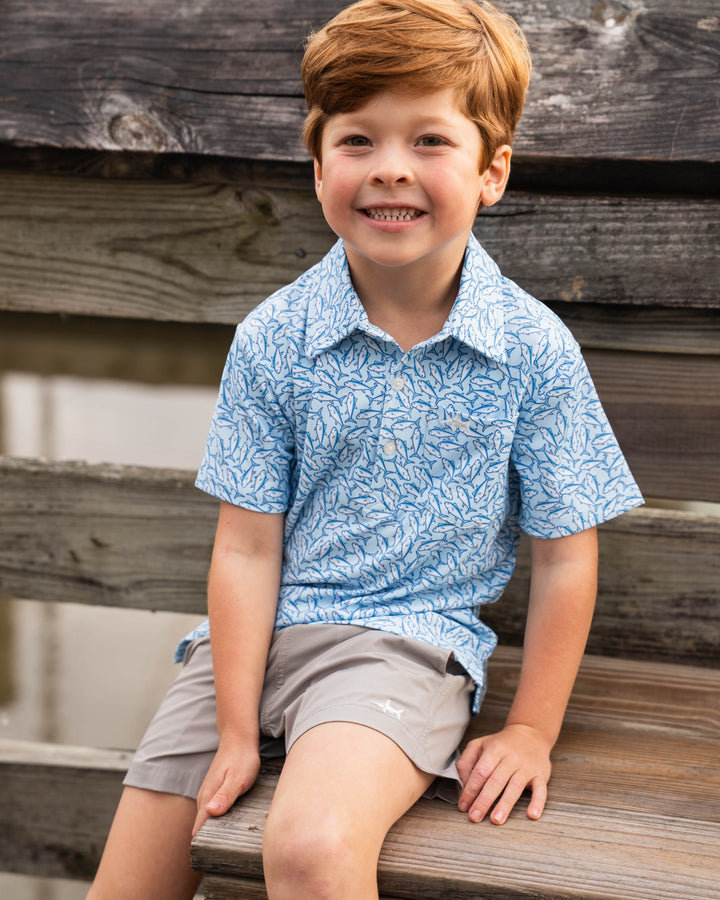Young boy sitting on a wooden bench wearing a blue patterned shirt and gray shorts.