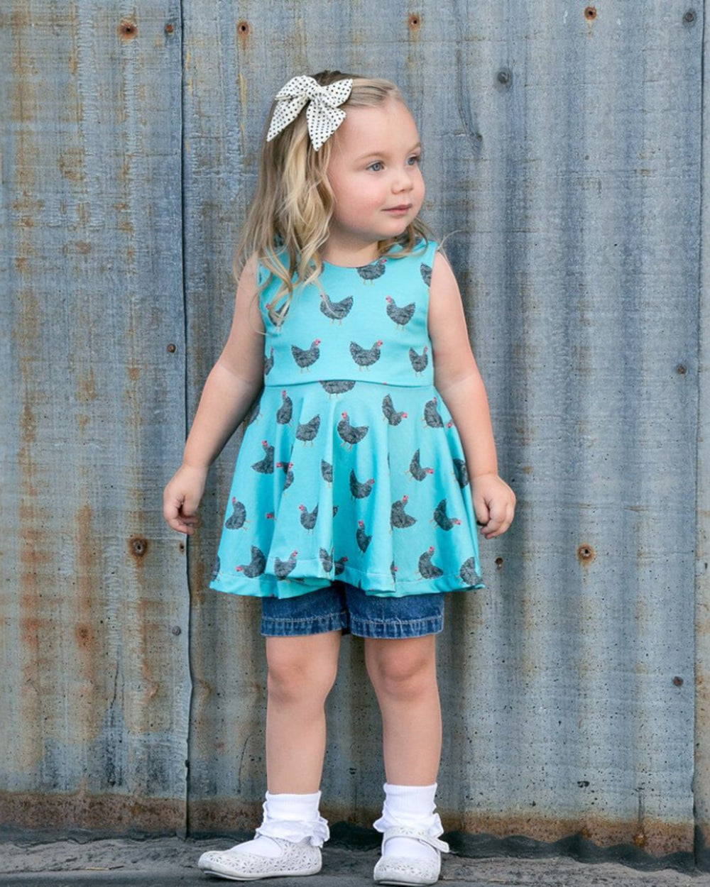 Young girl wearing a turquoise dress with chicken print and denim shorts against a rustic metal wall.
