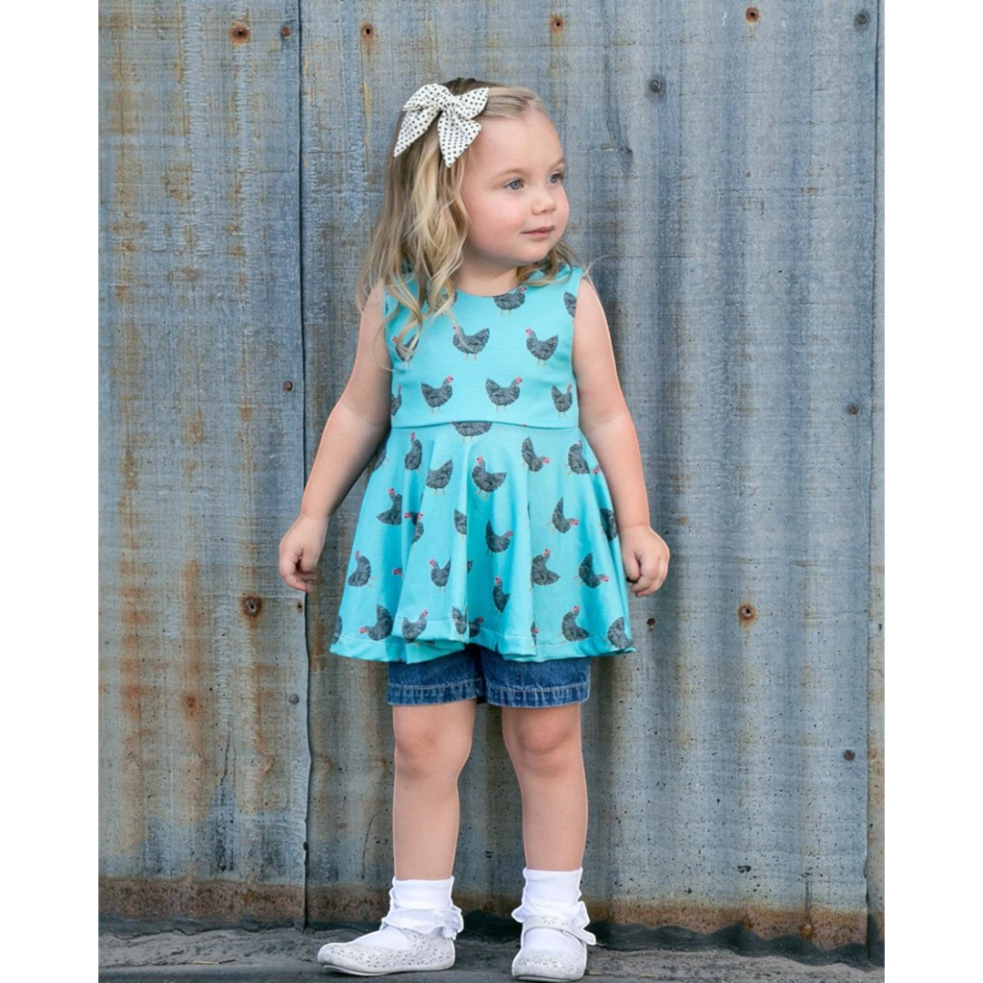 Young girl wearing a turquoise dress with chicken print and denim shorts against a rustic metal wall.