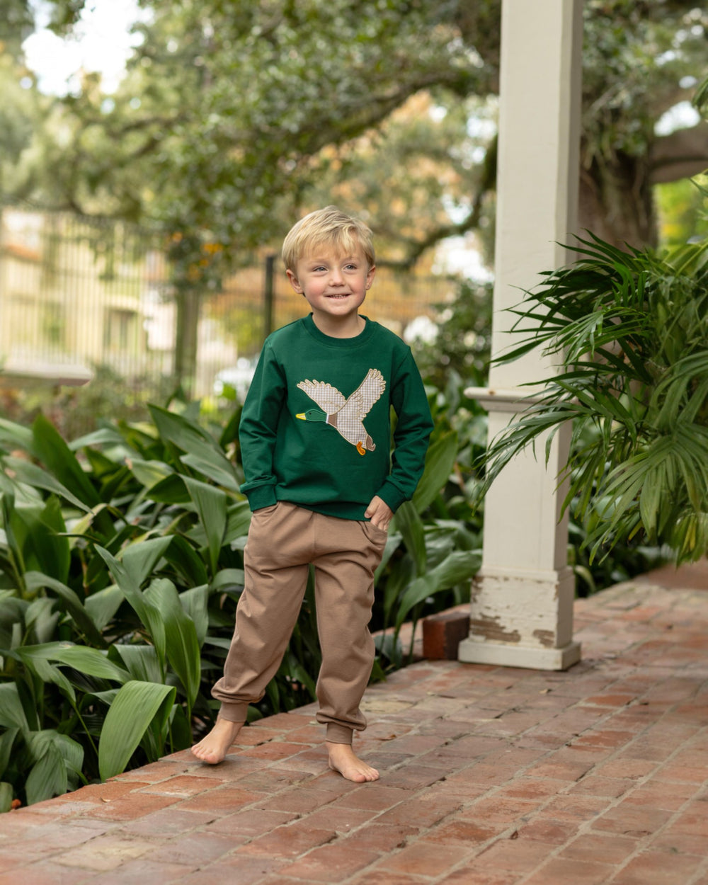 Young boy wearing a green sweater with a bird design, standing on a brick path surrounded by plants.
