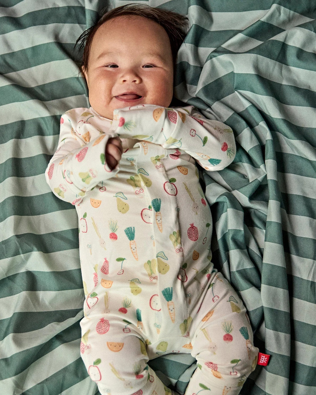 A baby lying on a green and white striped blanket, wearing a cream-colored footed sleeper with a colorful vegetable print. A small red tag with the brand name &