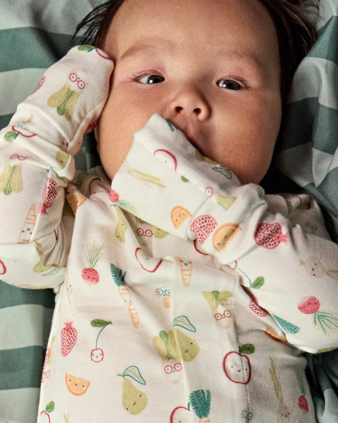 A close-up of a baby lying down wearing a long-sleeve cream-colored pajama top with a colorful fruit and vegetable print, set against a green and white striped blanket, that store