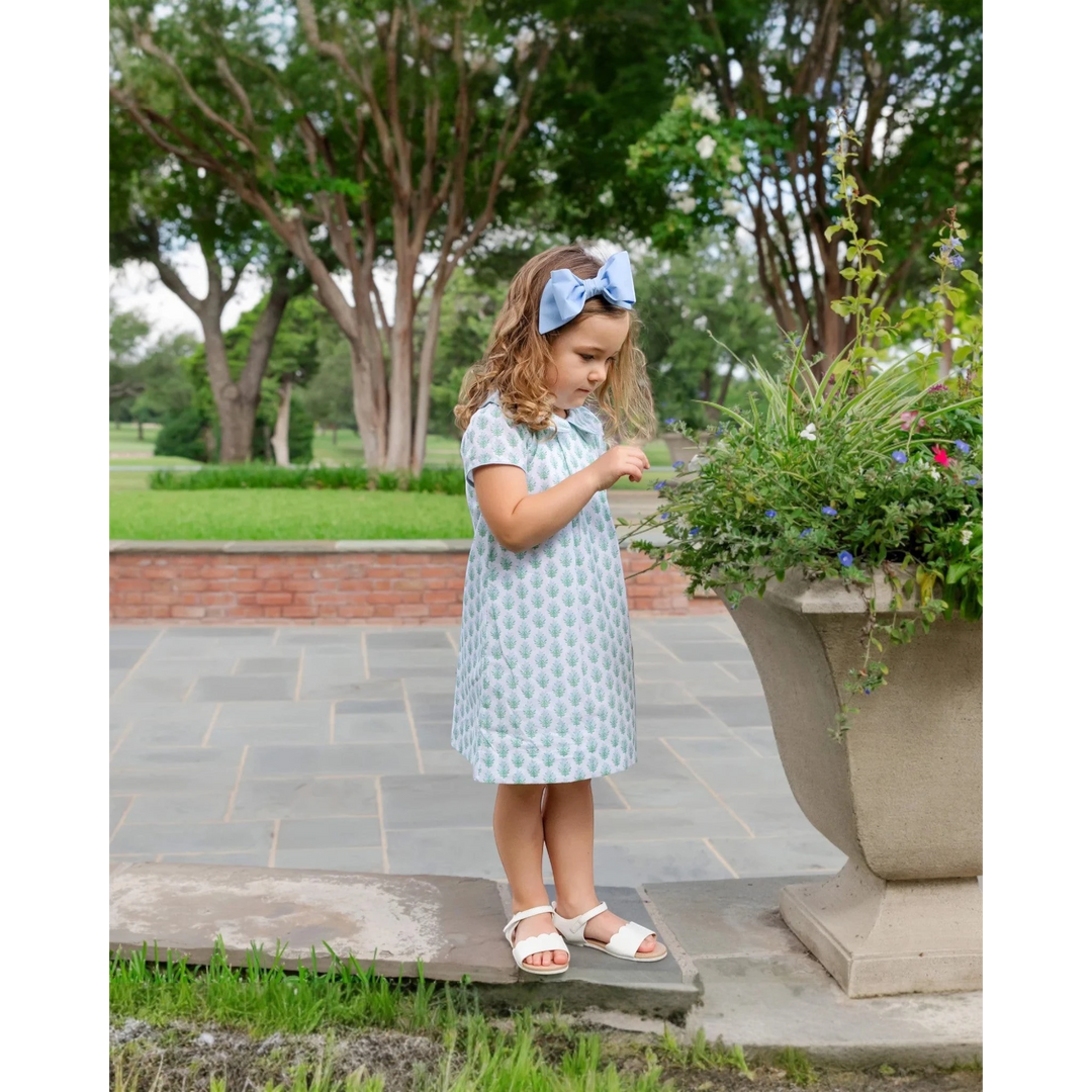 Young girl in a light blue dress standing outdoors near a planter with flowers.