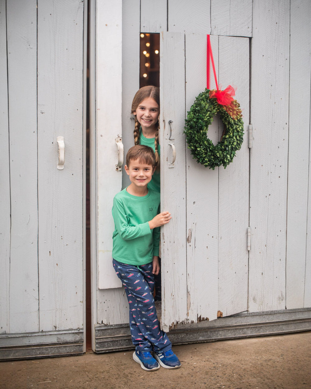 Two children standing in a doorway with a wreath hanging on a wooden door.
