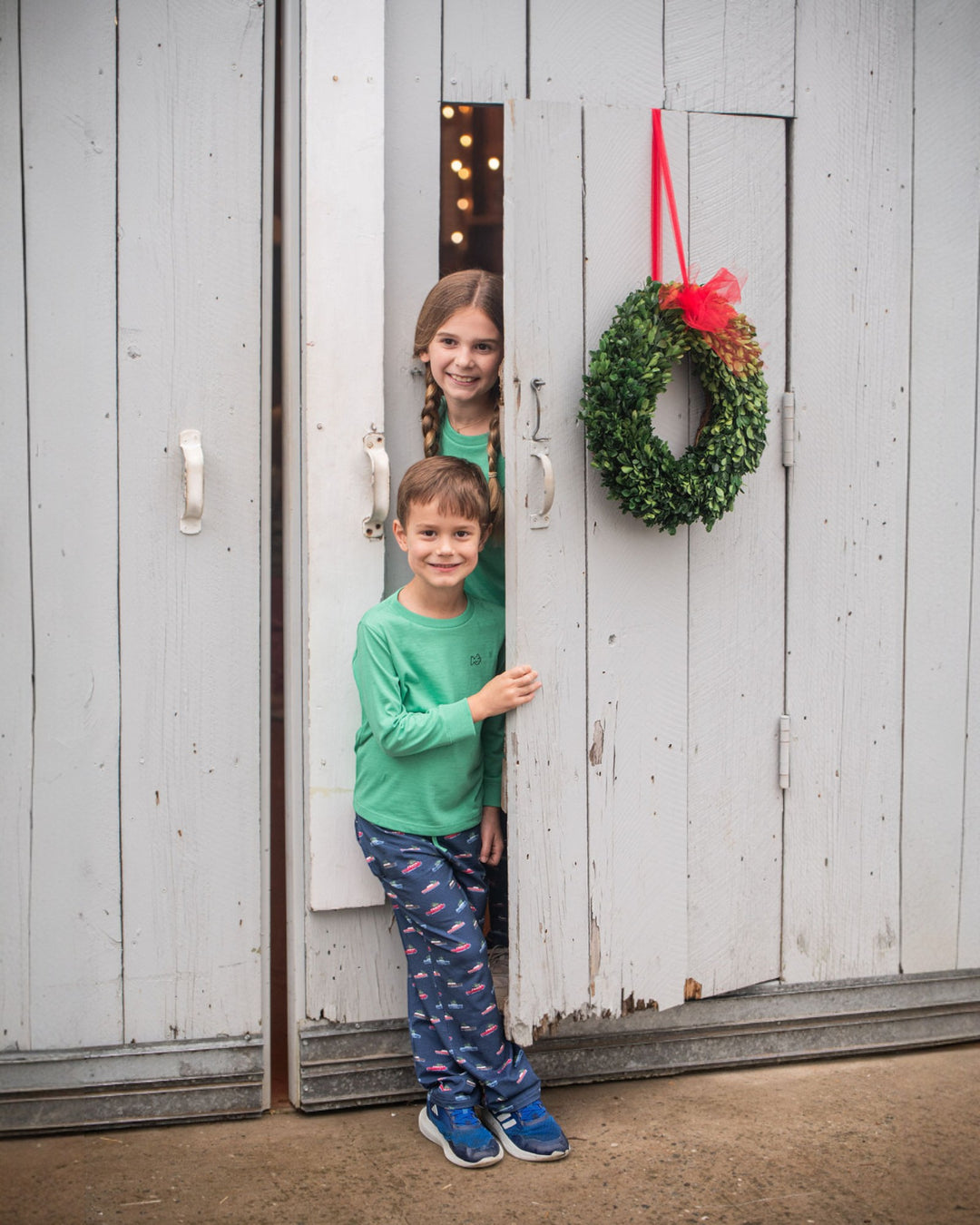 Two children standing in a doorway with a wreath hanging on a wooden door.