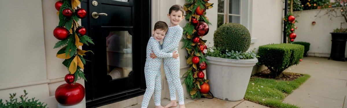 Two children hugging in front of a house door, wearing matching light blue pajamas with a white pattern. The scene is decorated with Christmas garlands, red ornaments, and potted plants at That Store