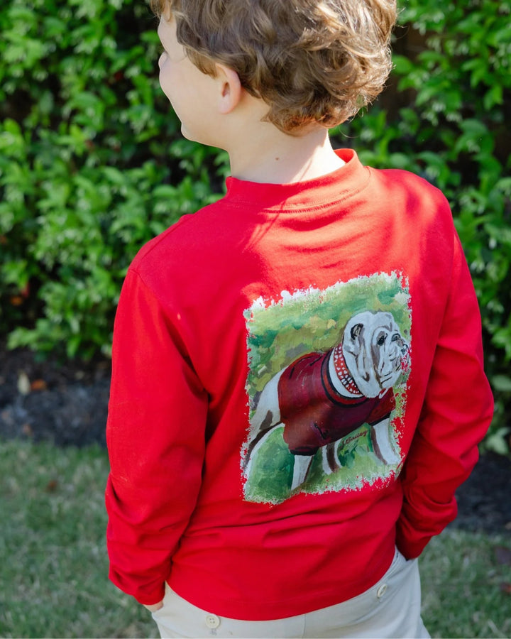 A child wearing a bright red long-sleeve shirt outdoors, shown from the back. The shirt features a large graphic print of a bulldog wearing a sweater on the back, that store
