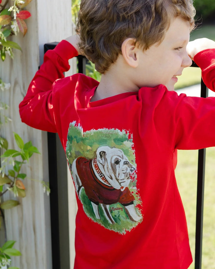 A child wearing a bright red long-sleeve shirt with a large printed graphic of an bulldog on the back, photographed outdoors near a fence and plants, that store