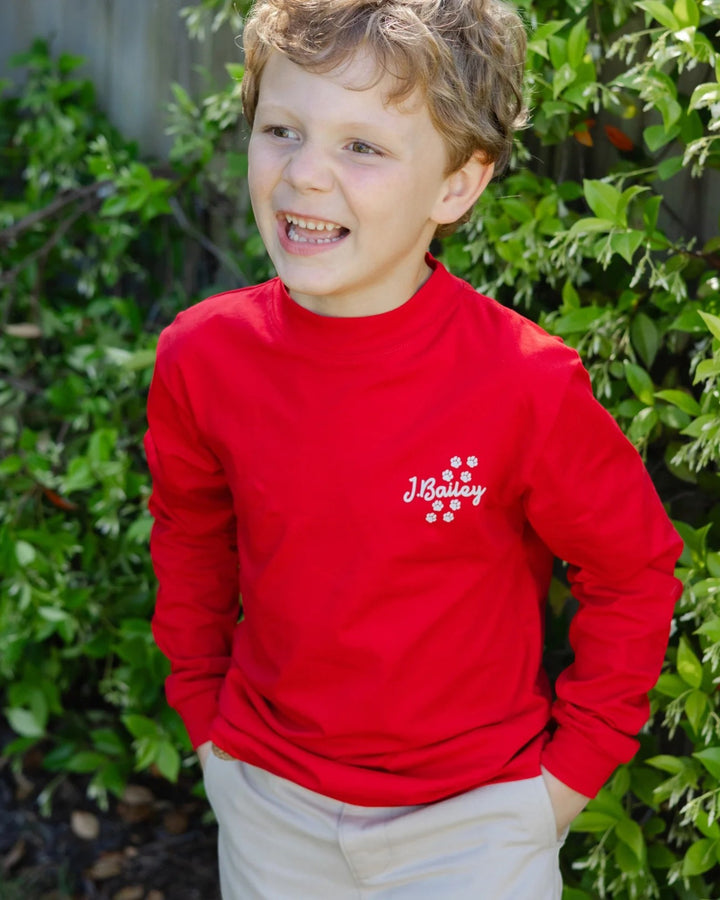 A young boy with curly light brown hair smiling outdoors, wearing a bright red long-sleeve mock neck shirt with &