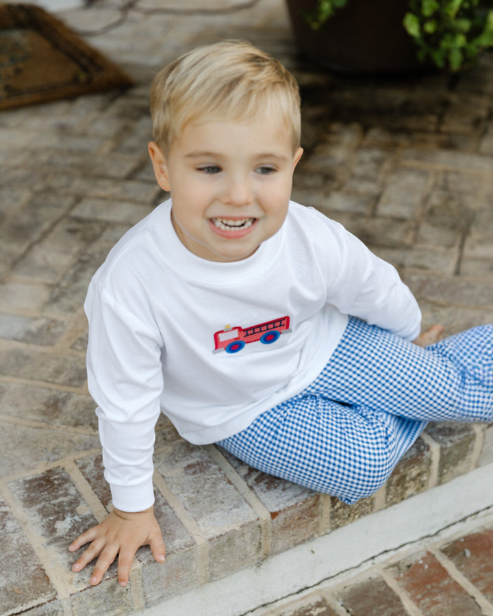 Child wearing a white long-sleeve shirt with a red fire truck design and blue checkered pants, sitting on a stone step.