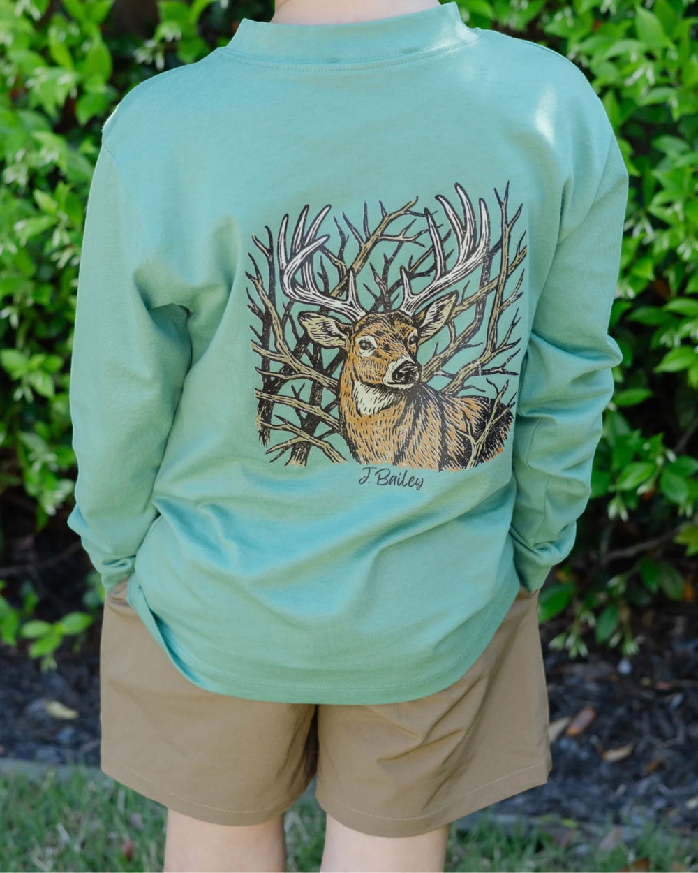 A person outdoors wearing a mint green long-sleeve shirt with a large graphic of a deer among branches on the back, paired with tan shorts, set against a background of green foliage, that store