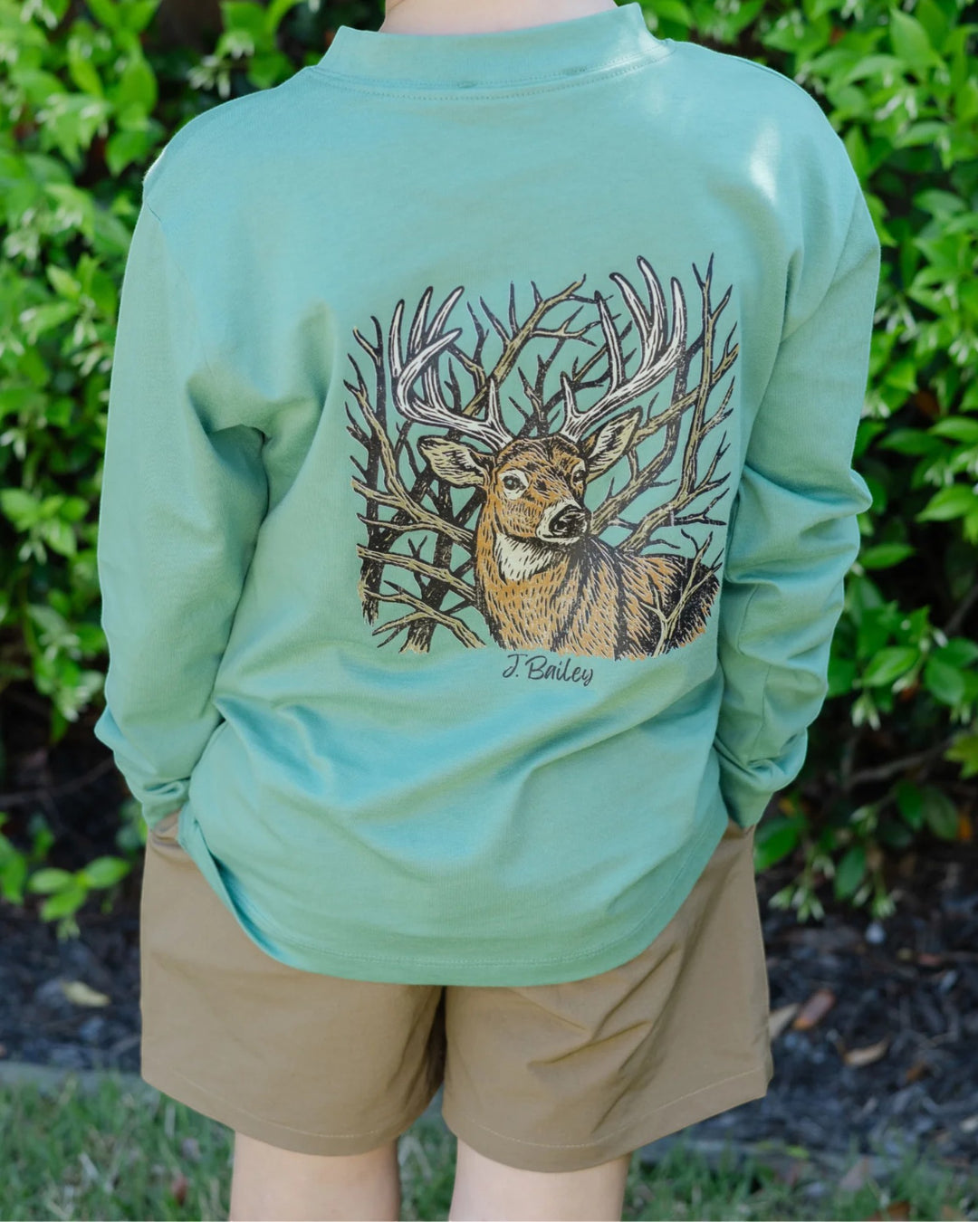 A person outdoors wearing a mint green long-sleeve shirt with a large graphic of a deer among branches on the back, paired with tan shorts, set against a background of green foliage, that store