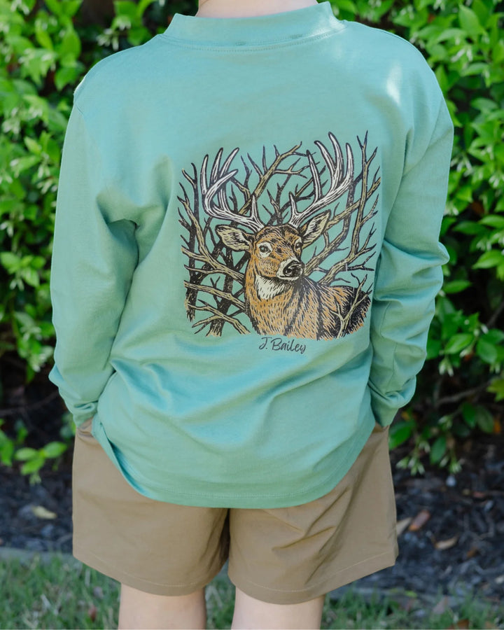 A person outdoors wearing a mint green long-sleeve shirt with a large graphic of a deer among branches on the back, paired with tan shorts, set against a background of green foliage, that store