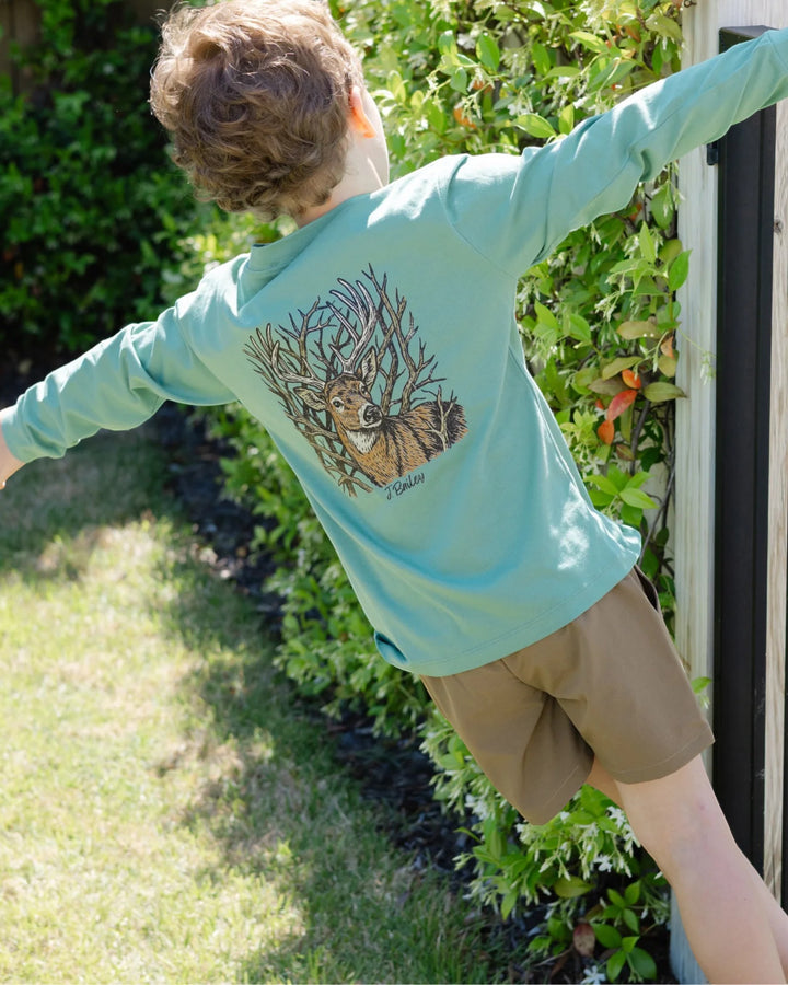 A child wearing a mint green long-sleeve shirt with a large deer graphic on the back, paired with tan shorts, posing outdoors in a garden setting, that store