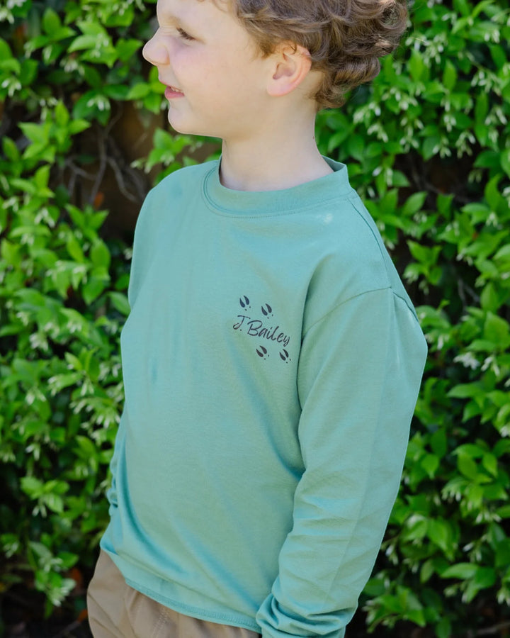 A child wearing a sage green long-sleeve crewneck sweatshirt with a black embroidered &