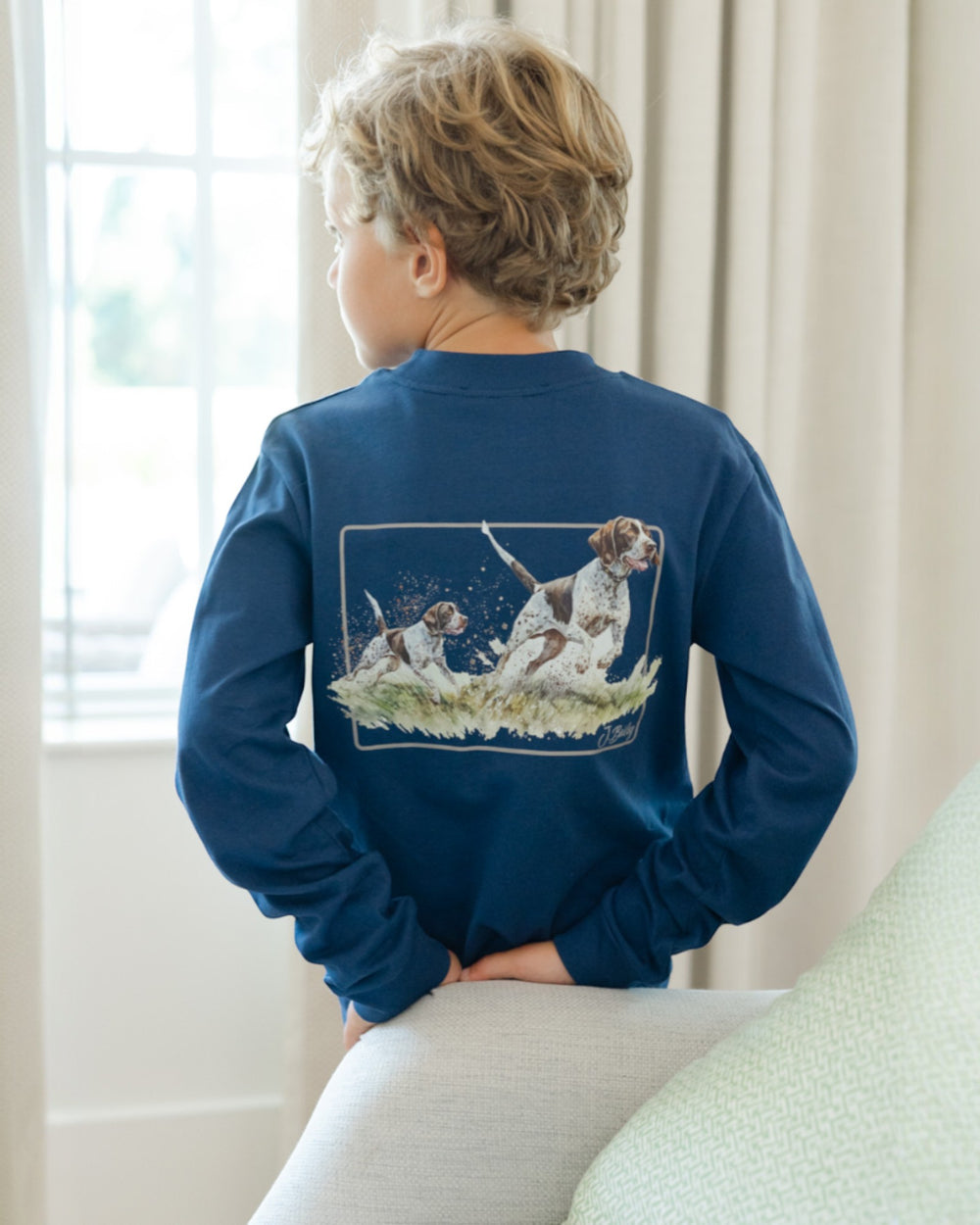 Child wearing a blue long-sleeve shirt with a graphic of dogs on the back, sitting in a room with light-colored curtains.