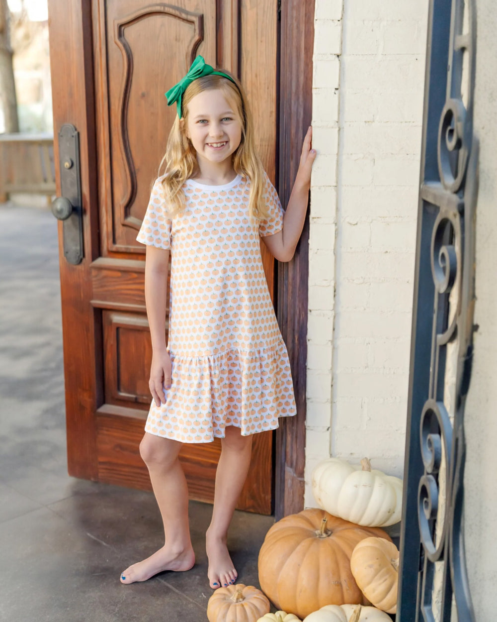 Young girl in a polka dot dress standing next to pumpkins in front of a wooden door.