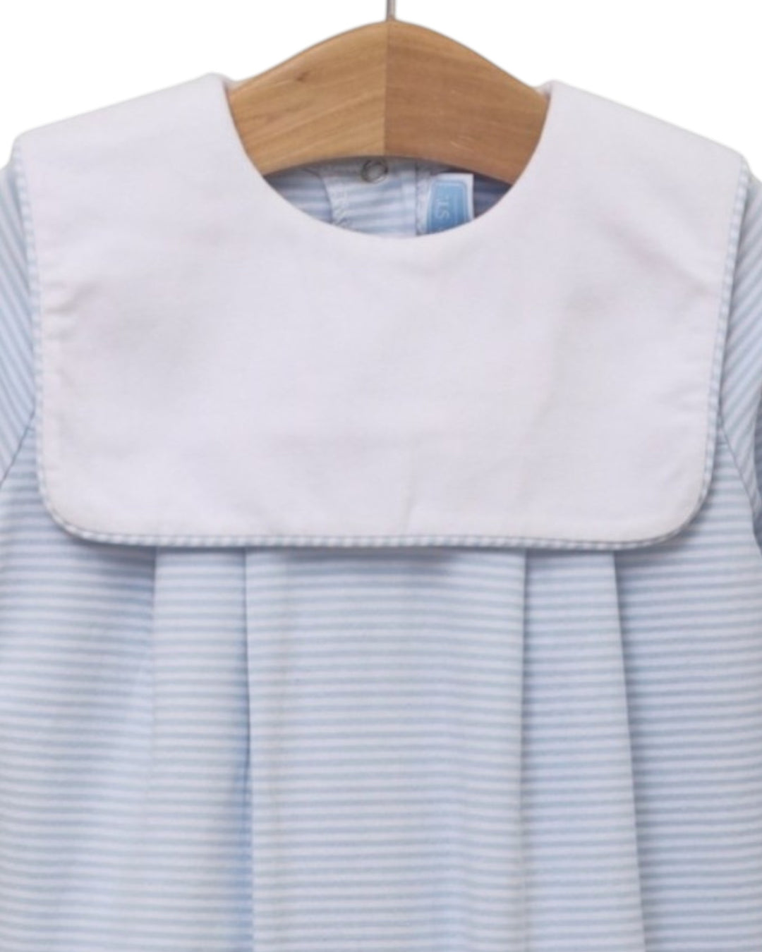 A close-up of a baby dress or romper with a wide white bib collar and light blue and white horizontal stripes, hanging on a wooden hanger against a white background, that store