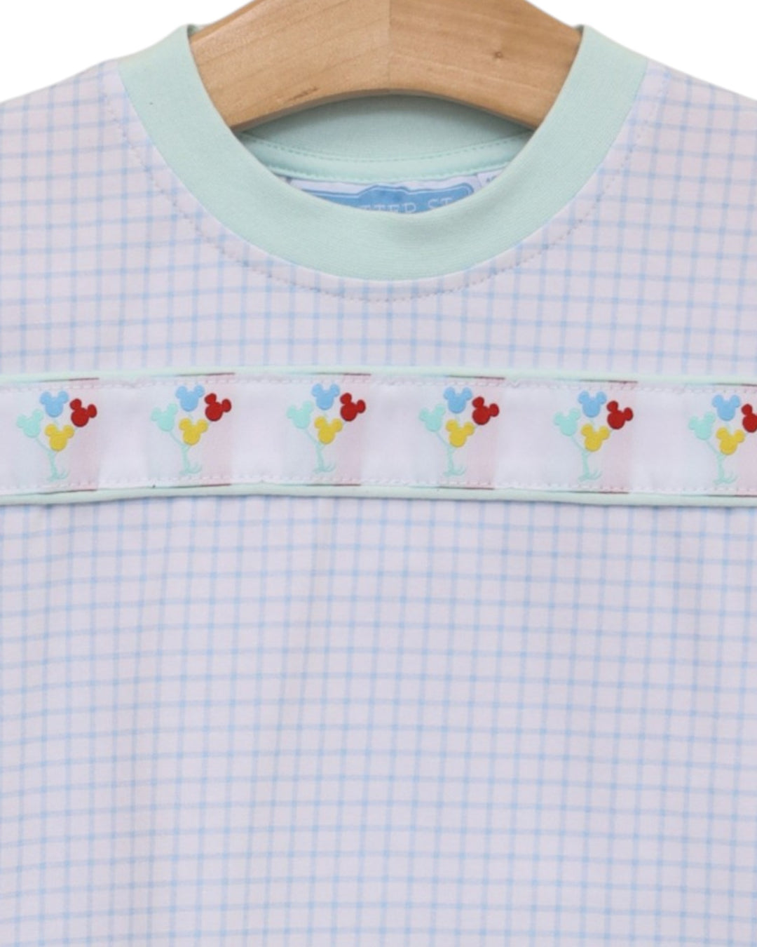 A close-up of a baby or toddler long-sleeve top on a wooden hanger, featuring a light blue and white gingham pattern, a mint green crew neck, and a decorative horizontal band with colorful embroidered flowers and hearts, that store