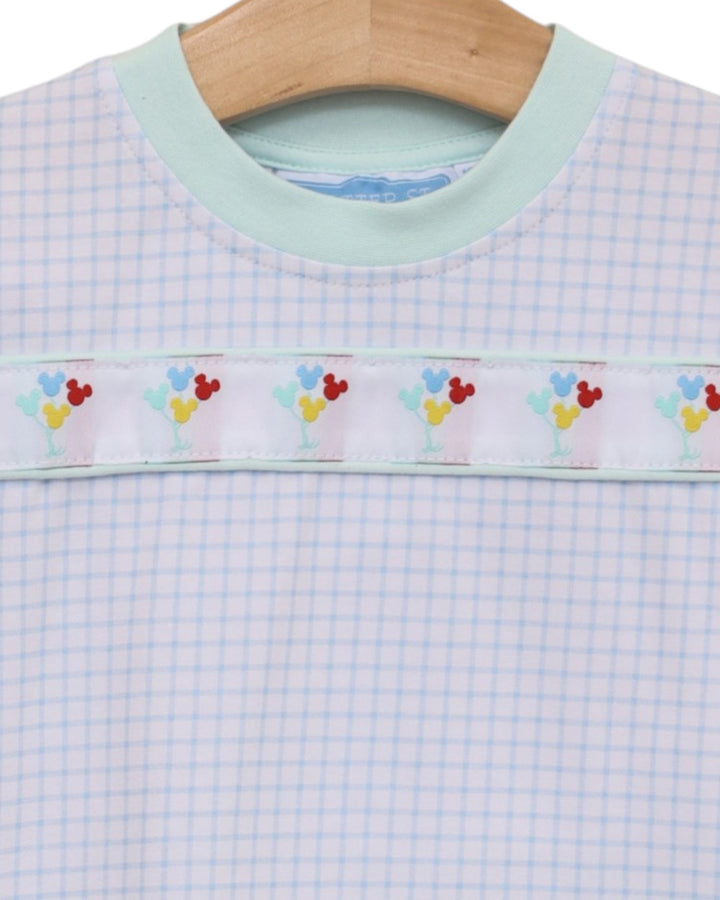 A close-up of a baby or toddler long-sleeve top on a wooden hanger, featuring a light blue and white gingham pattern, a mint green crew neck, and a decorative horizontal band with colorful embroidered flowers and hearts, that store