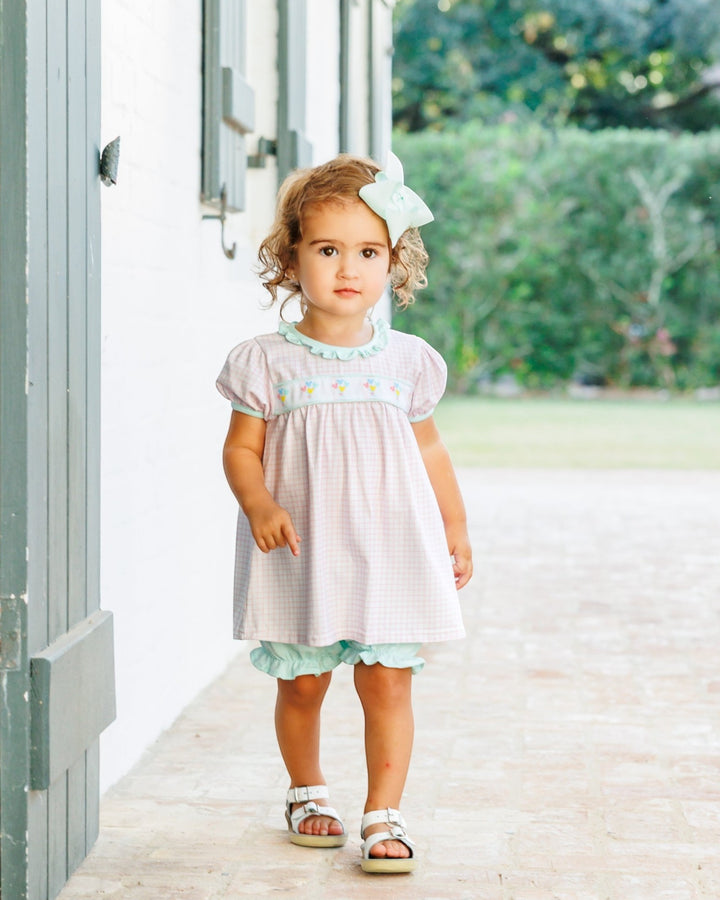 A toddler girl wearing a light pink and white gingham smocked dress with mint green ruffle trim, a matching bow in her hair, and sandals, standing on a stone patio next to a white wall, that store