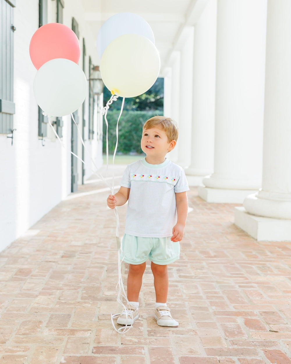 A toddler stands on a brick porch holding a string of pastel balloons, wearing a white short-sleeve top with colorful embroidered details and mint green shorts, that store