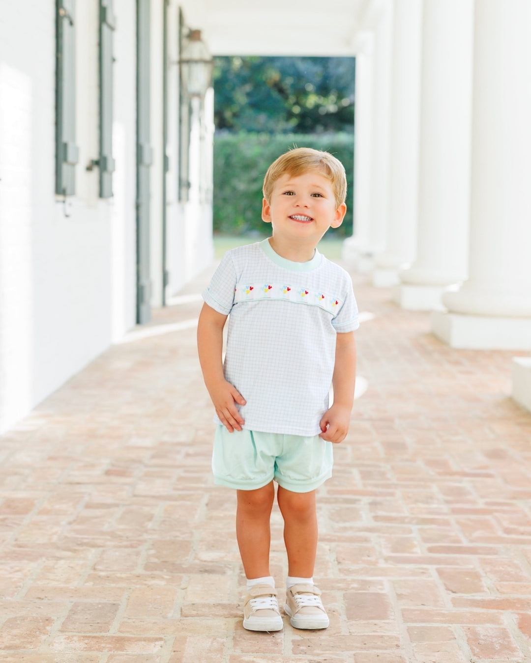 A young boy stands on a brick walkway in front of a white building with columns, wearing a white short-sleeve t-shirt with a small red embroidered pattern across the chest and light mint green shorts, that store